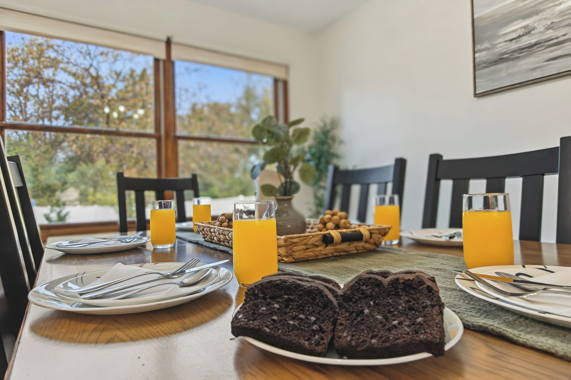 Ein Frühstückstisch mit Stühlen, Tellern, Besteck, Gläsern Orangensaft und einem Schokoladenkuchen.