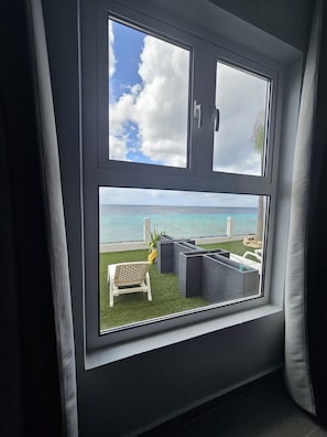 Una habitación con un amplio ventanal con vista a una playa, cielo despejado, zona verde y mobiliario exterior.