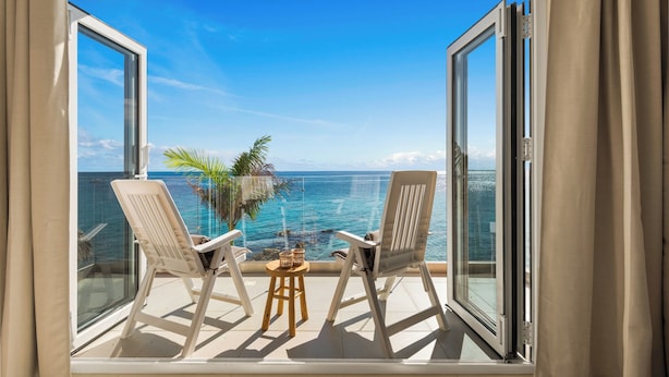 A balcony with two chairs and a small table, overlooking a clear blue sea and a palm tree. - Seaview Resort Boca Sami (Sint Michiel)