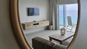 A modern hotel room with a large oval mirror, a wooden TV stand, a sofa, a dining table, and a view of the sea through large windows. - Seaview Resort Boca Sami (Sint Michiel)
