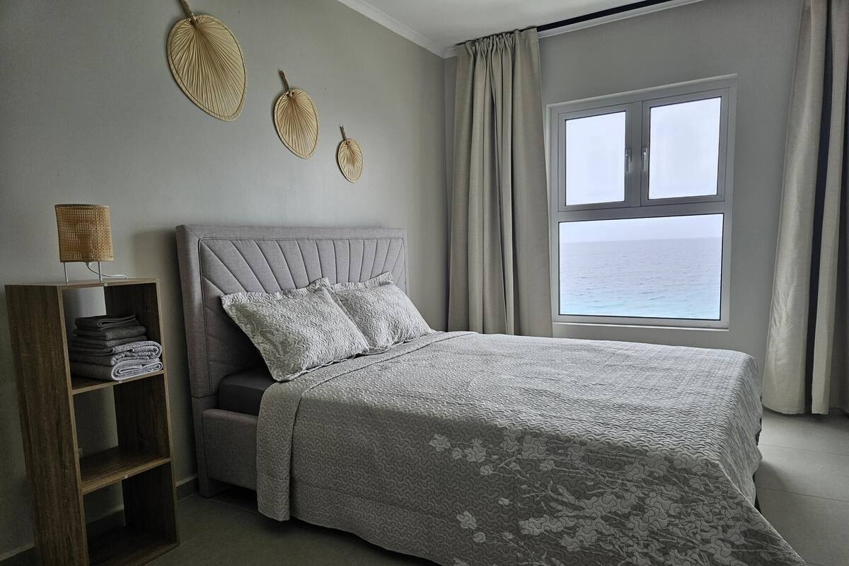 Quarto com cama, mesa de cabeceira, janela com vista para o mar e decoração na parede.