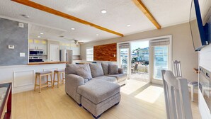 Una sala de estar moderna con un sofá modular gris, vigas de madera en el techo y una cocina con gabinetes blancos y horno empotrado.