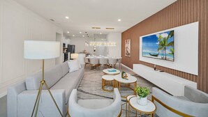 Una sala moderna con un sofá, sillones, una mesa de centro, una lámpara de pie, un comedor con mesa y sillas, y un televisor montado en la pared.