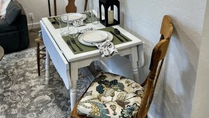 A dining area with a table set for two, a chair with a floral cushion, a black lantern, and two framed floral paintings on the wall. - Cozy cottage in downtown Hamilton in the heart of the Bitterroot Valley.  (Hamilton)