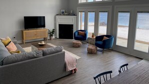 A spacious living room with a large window overlooking a beach, a fireplace, and a television on a wooden cabinet. - Lake front living with private sandy beach! (Elizabethtown)