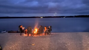 A group of people seated around a campfire on a sandy beach with a calm lake in the background. - Lake front living with private sandy beach! (Elizabethtown)