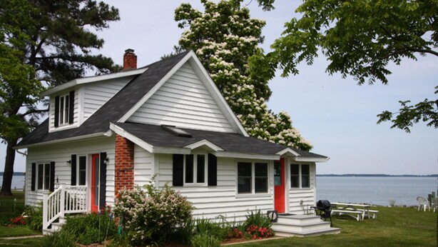 A white house with a red door and chimney, surrounded by greenery and a view of the water. -  Artists, writers & creative minds  IMAGINE YOUR GETAWAY AT SIMPLICITY COTTAGE (Tilghman Island)