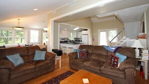 A spacious living room with two brown sofas, a wooden coffee table, and a kitchen in the background. -  Artists, writers & creative minds  IMAGINE YOUR GETAWAY AT SIMPLICITY COTTAGE (Tilghman Island)