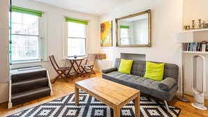 Un salon avec un canapé gris, une table basse en bois et un tapis à motifs. On y trouve également un coin repas avec une table et des chaises, ainsi qu’un grand miroir accroché au mur.