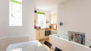 Une cuisine compacte avec un coin repas, comprenant une table blanche, une fenêtre avec un store vert et une vue sur la rue.