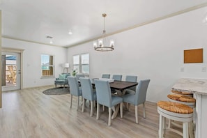 Uma sala de jantar com uma mesa redonda, seis cadeiras, um lustre e vista para o exterior.