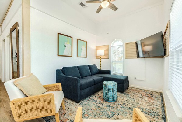 A living room with a sectional sofa, a rattan chair, a round ottoman, a patterned rug, a ceiling fan, and a mounted TV. - Steps to Sand Renovated Surfside Beach House (Surfside Beach)