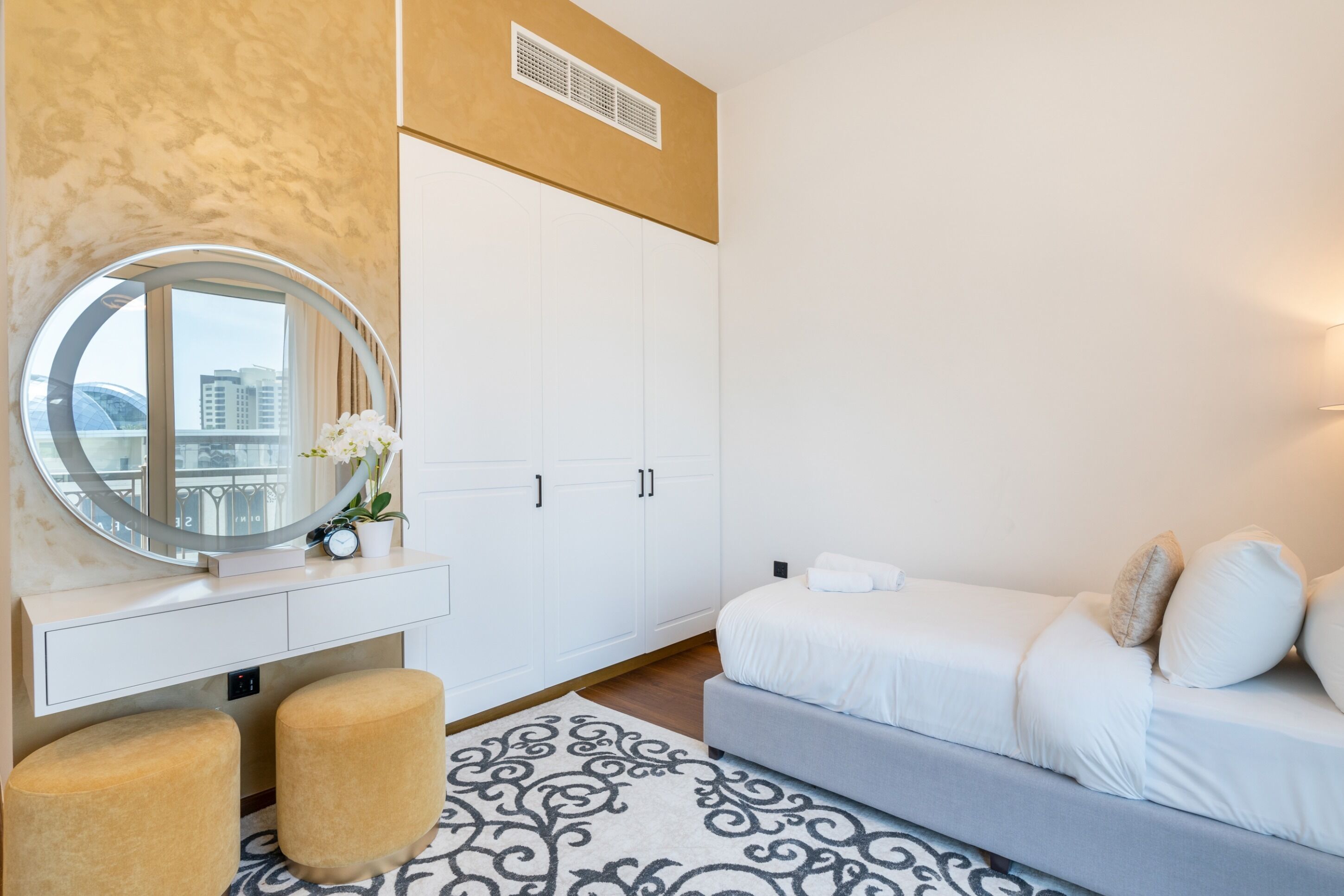 Ein modernes Hotelzimmer mit einem großen Spiegel, einer weißen Kommode, zwei Poufs, einem grauen Sofa und einem gemusterten Teppich.