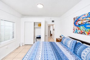 A bedroom with a bed, a window with blinds, a door, a hallway, and a colorful abstract painting on the wall. - Route 66 Downtown Blue Bungalow w/Fenced Yard, AC, W/D, 4 Smart TVs, Fast Wi-Fi (Williams)