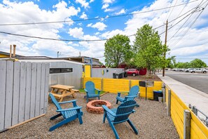 A fenced outdoor patio with wooden furniture, a fire pit, and a yellow wall. - Route 66 Downtown Blue Bungalow w/Fenced Yard, AC, W/D, 4 Smart TVs, Fast Wi-Fi (Williams)