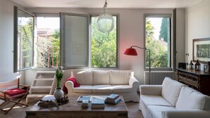 A spacious living room with a white sofa, a coffee table with books, a red floor lamp, and large windows offering a view of greenery outside. - Canal Views and a Secret Garden in Giudecca (Venezia)