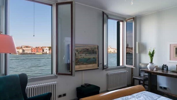 A hotel room with a bed, a desk, a chair, and a view of the water and buildings. - Canal Views and a Secret Garden in Giudecca (Venezia)