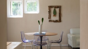 A dining area with a round table, four transparent chairs, a white vase with a cactus, and two windows with a view of trees. - Cozy House close to the sea (Foxi Lioni)