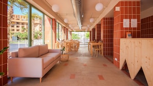 A modern hotel lobby with a sofa, dining area, and large windows offering views of the outside. - Aparthotel Terra Aurea (Salou)