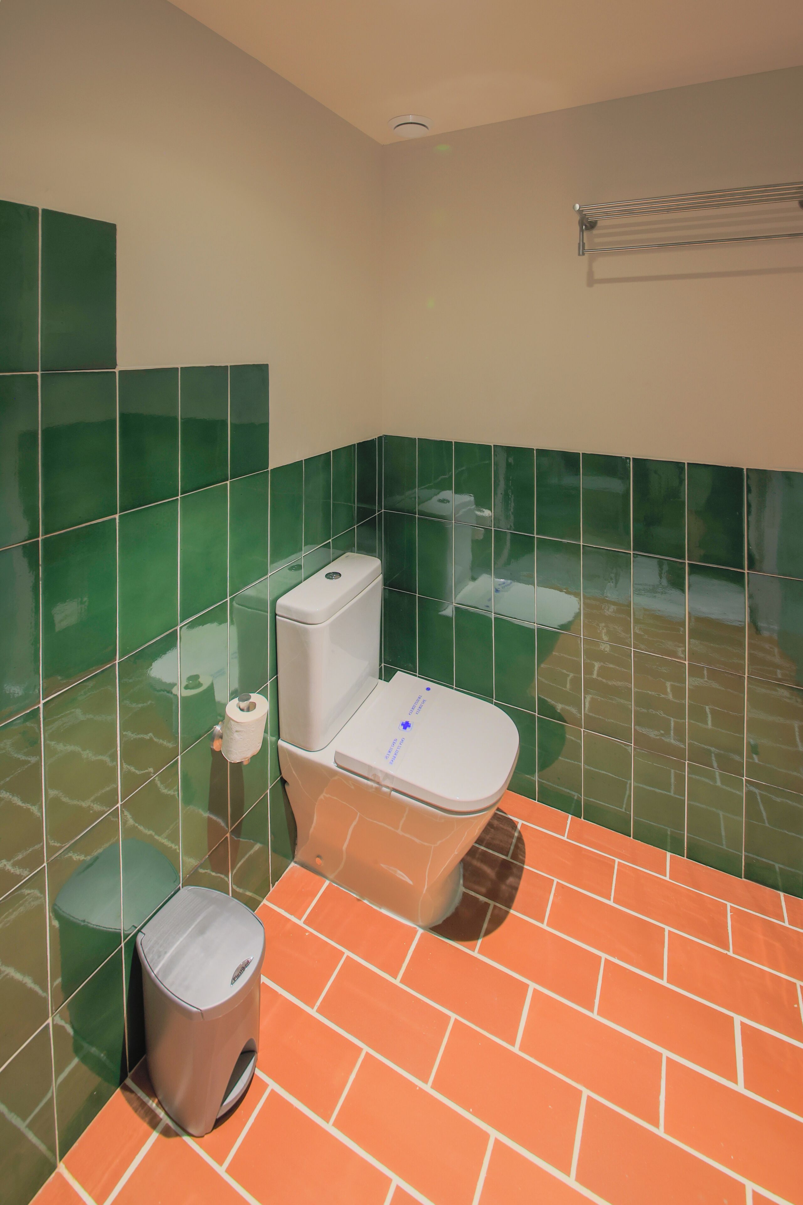 Een badkamer met wandtegels in groen en oranje, een wit toilet, een prullenbak en een handdoekhouder.