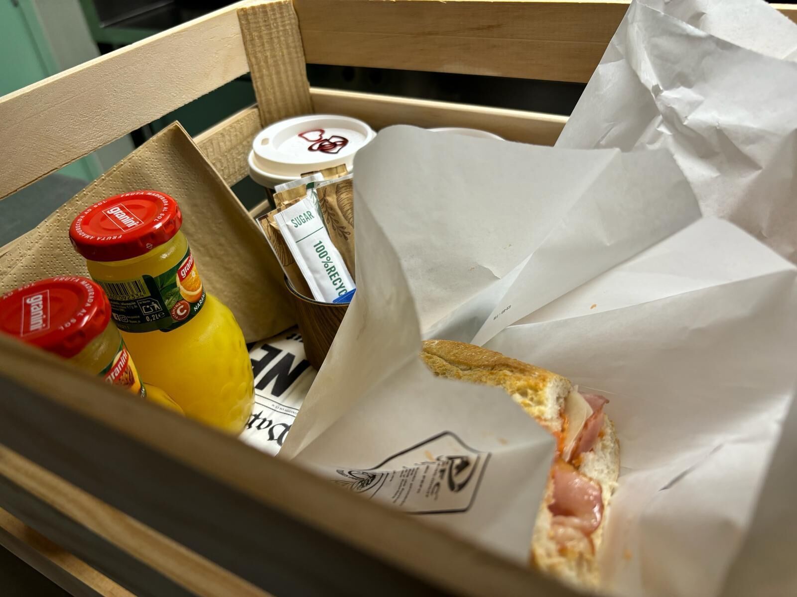 Una caja de madera con un sándwich, una botella de mostaza y un sobre de azúcar.