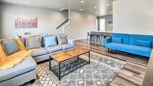 Ein modernes Wohnzimmer mit grauem Sofa, blauem Sofa und Couchtisch aus Holz. Eine Treppe führt zu einer weiteren Ebene, und an der Wand hängt ein farbenfrohes abstraktes Gemälde.