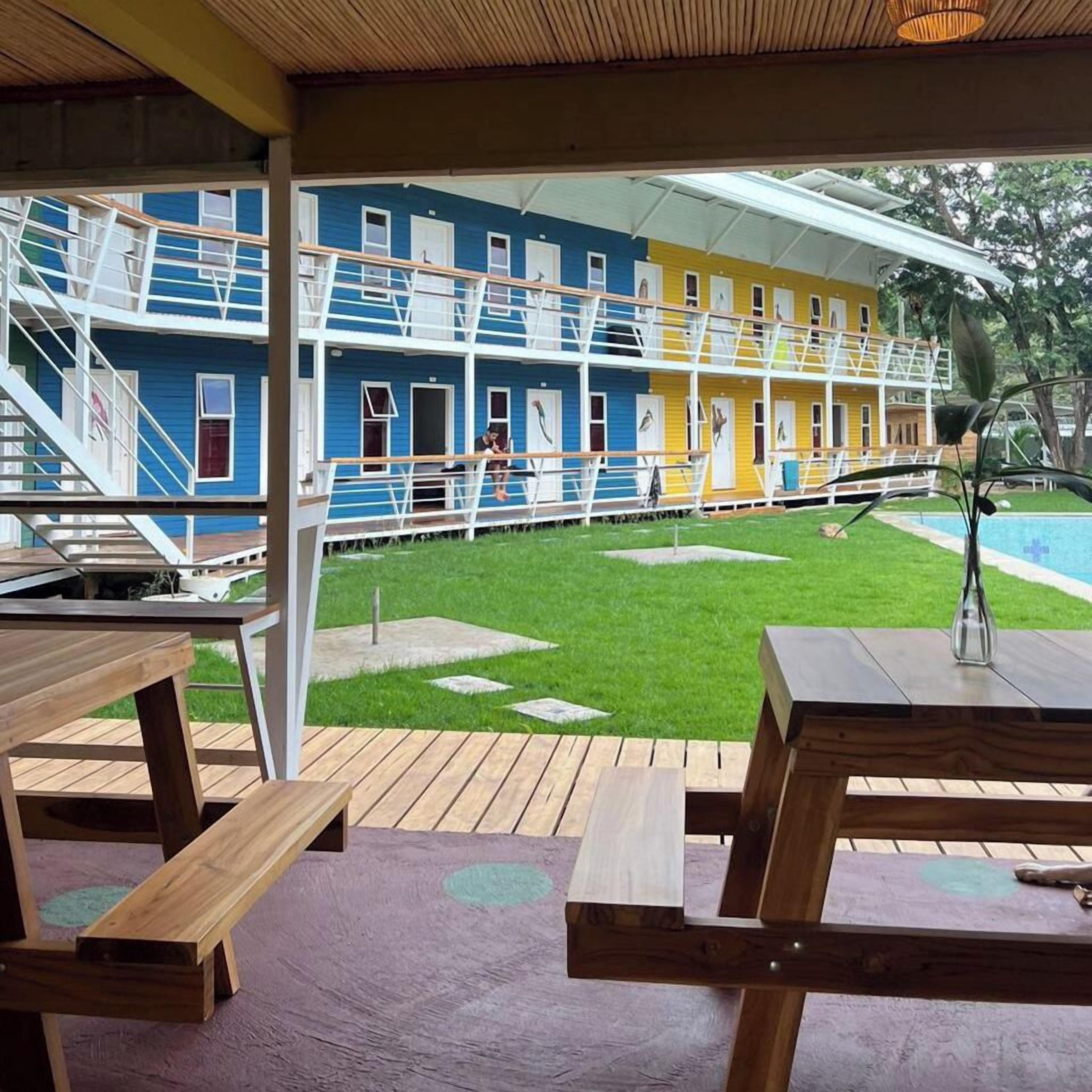 A colorful building with a covered outdoor area, wooden picnic tables, and a grassy lawn.