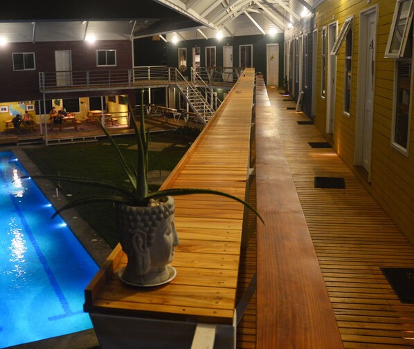 A wooden deck with a potted plant, a pool, and a building with multiple doors and windows in the background.