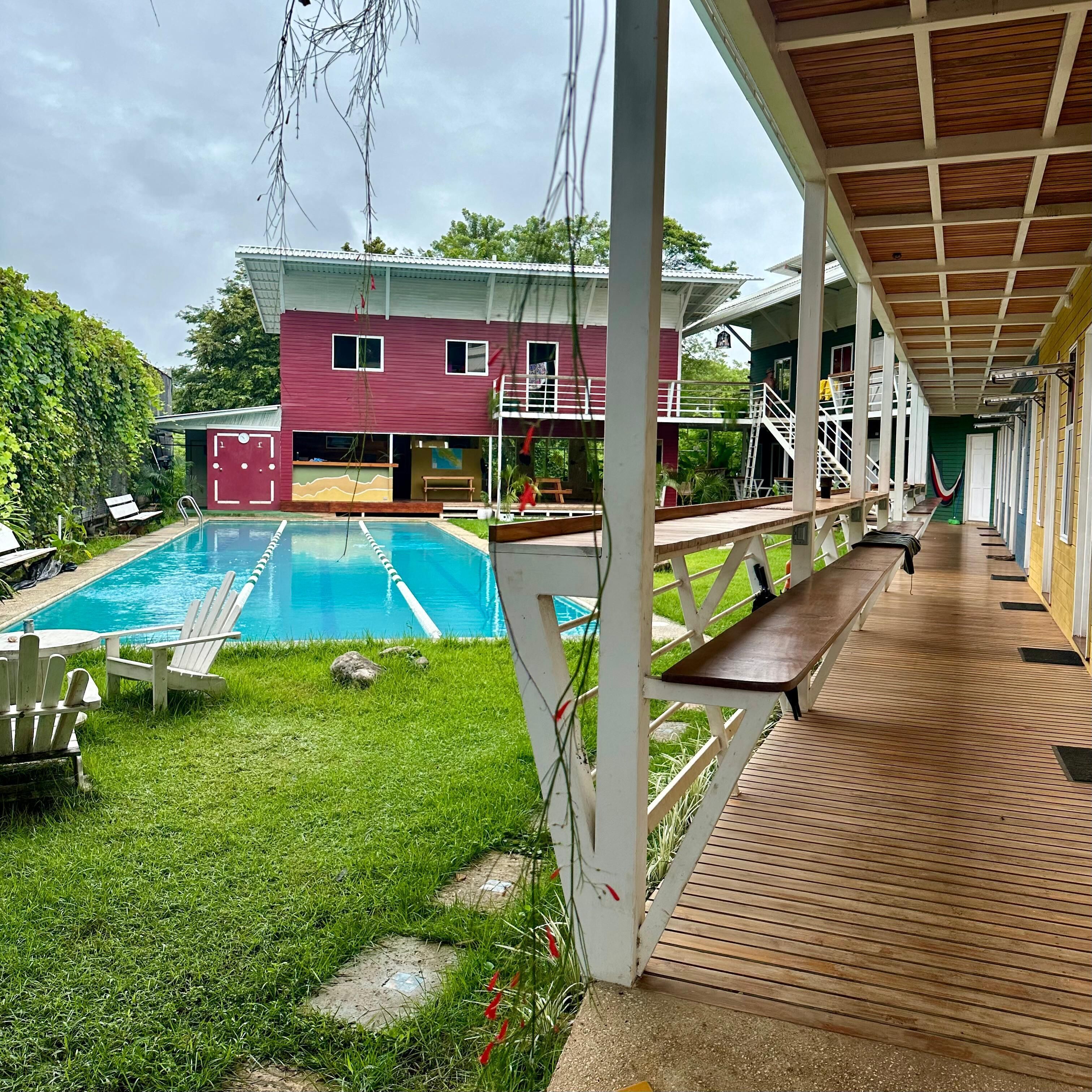 A covered pool area with a wooden deck, a red building in the background, and a clear blue pool.
