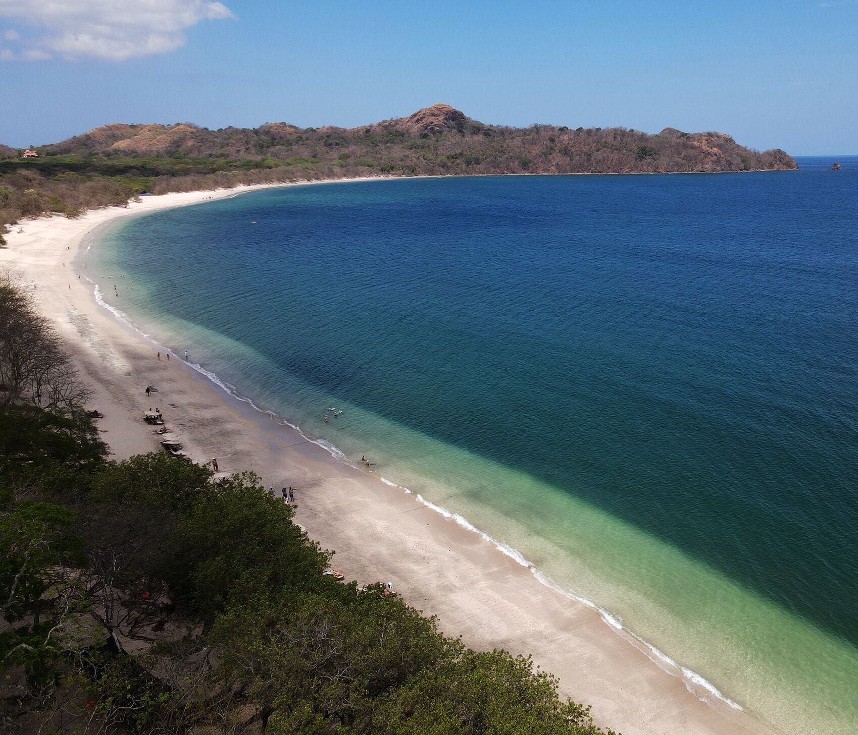 A beach with clear turquoise waters, a sandy shore, and a forested hill in the background.