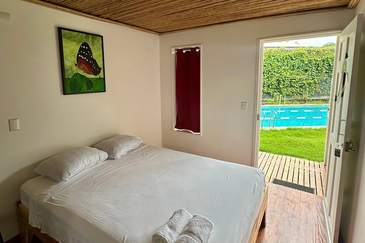 Un dormitorio con cama, un cuadro, una ventana con cortina y vista a una piscina.