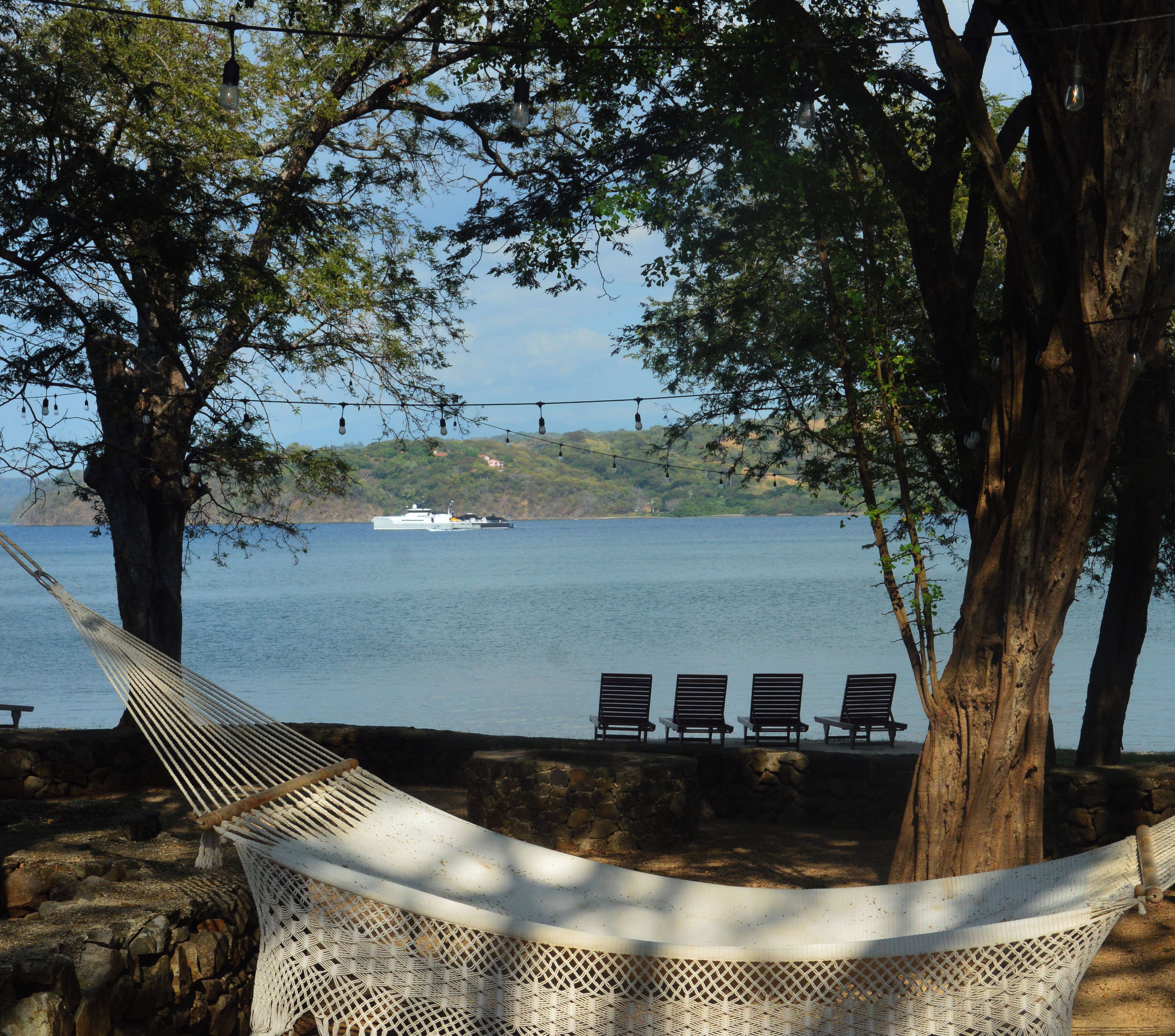 A hammock tied between two trees overlooking a body of water with a boat in the distance and a row of chairs along the shore.