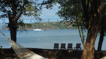 A hammock tied between two trees overlooking a body of water with a boat in the distance and a row of chairs along the shore.
