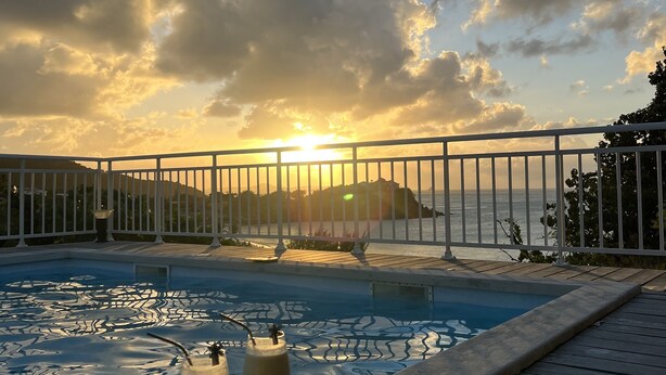 Une piscine avec deux verres d’une boisson, une terrasse en bois et une vue sur le coucher de soleil sur l’eau.