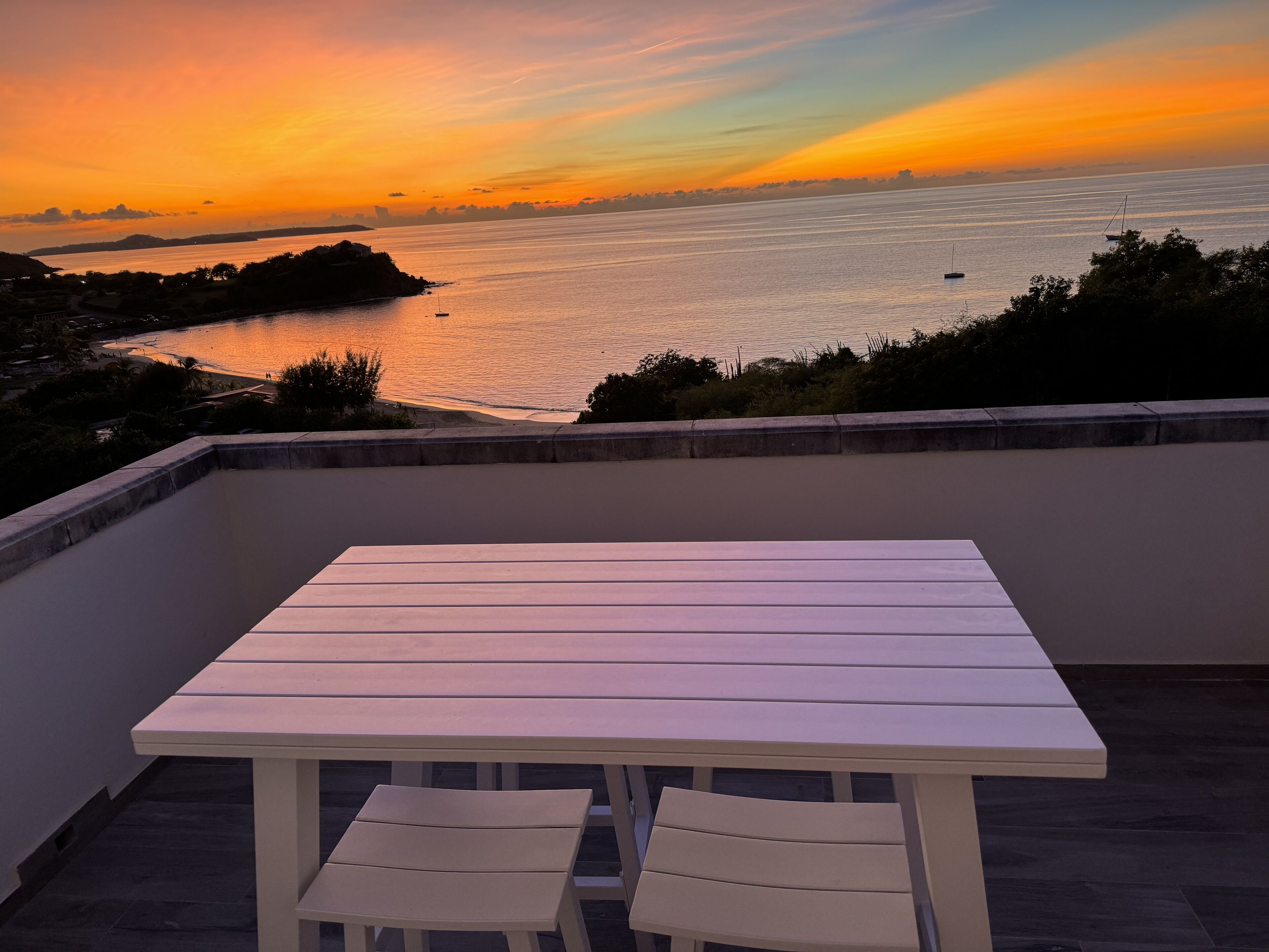 A terrace with a white table and two chairs, overlooking a body of water with boats and a sunset.