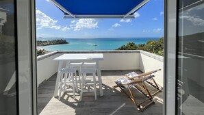 Un balcon avec une table et des chaises blanches, une chaise longue et une vue sur la mer et une petite île.
