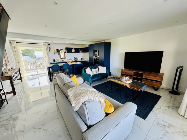 Een moderne woonkamer met een grijze bank, een blauwe fauteuil en een salontafel. Er is een keuken met donkere kasten en hanglampen. Een grote flatscreen televisie is aan de muur bevestigd.