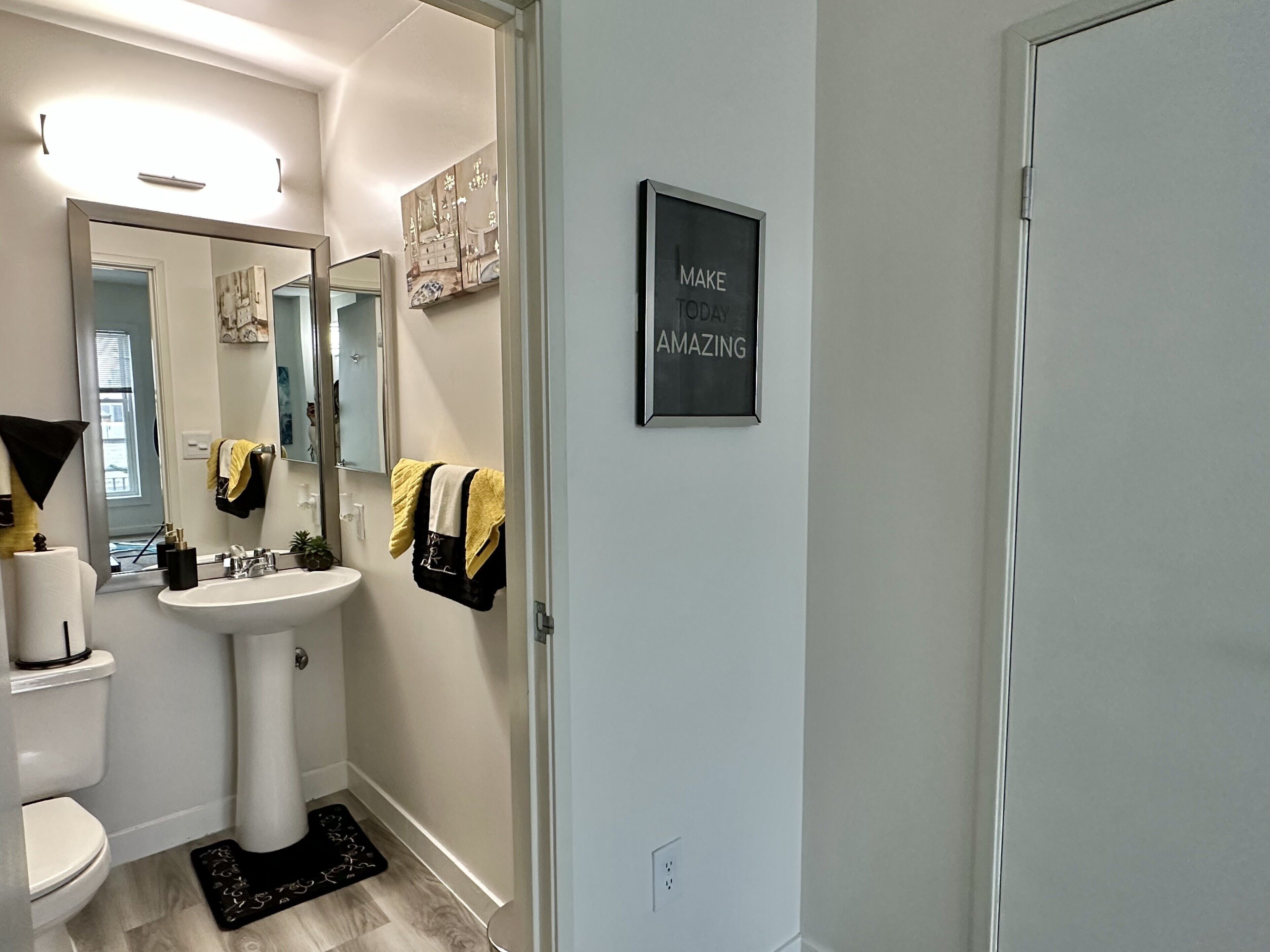 Une salle de bain moderne avec un lavabo sur colonne, des toilettes, un grand miroir et une citation inspirante sur le mur.