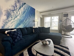 Ein modernes Wohnzimmer mit einem dunkelblauen Ecksofa, einem runden Couchtisch mit Dekoration, einem hängenden Eierstuhl und einem großen abstrakten Gemälde an der Wand.