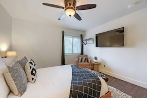 A bedroom with a bed, pillows, a ceiling fan, a mounted TV, and a window with blinds.