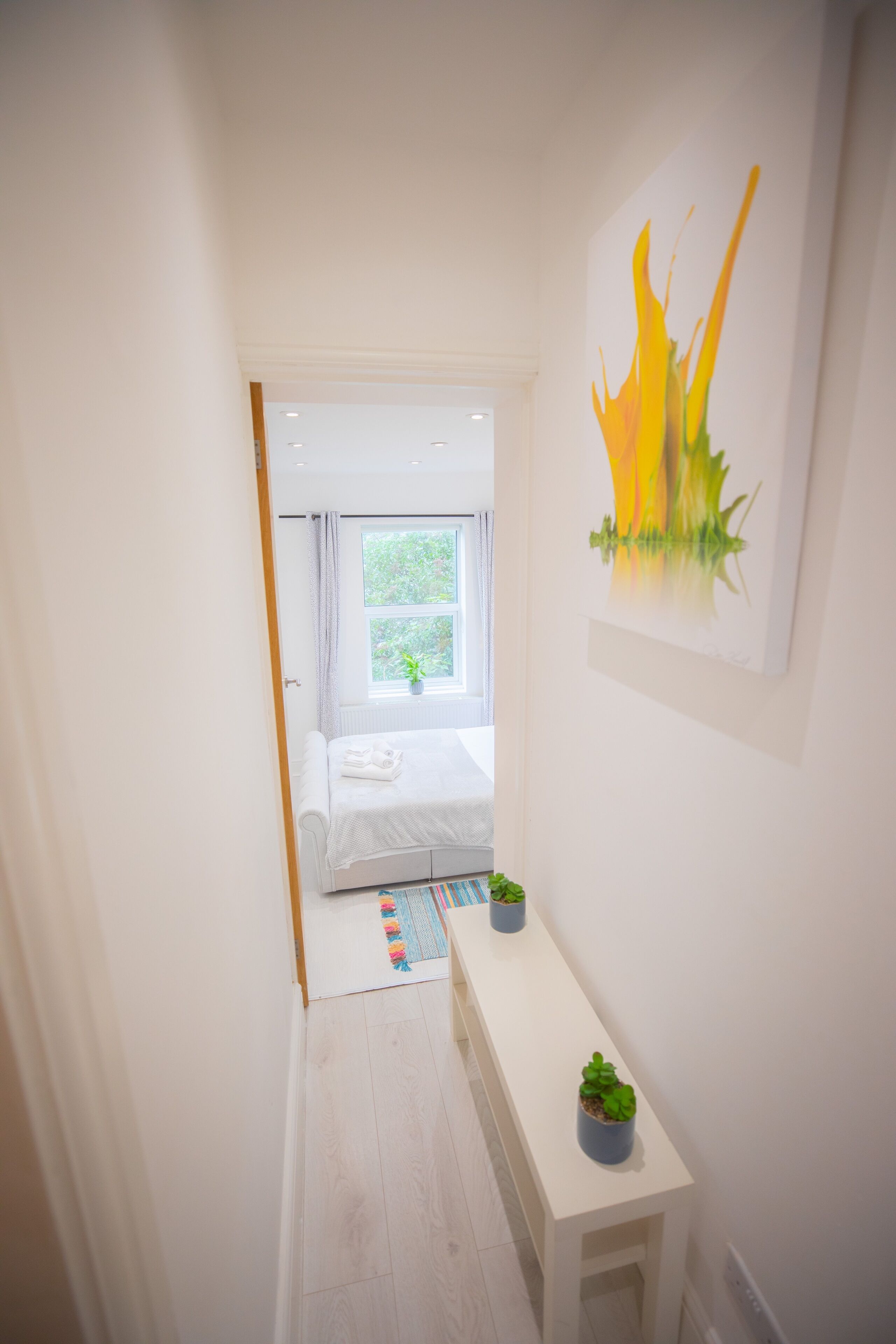 A narrow hallway leading to a bedroom with a bed, a window, and a painting on the wall.