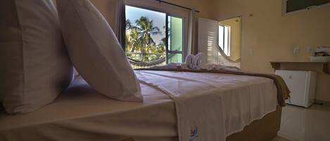A hotel room with a bed, a small desk, a window with a view of palm trees, and a hammock.