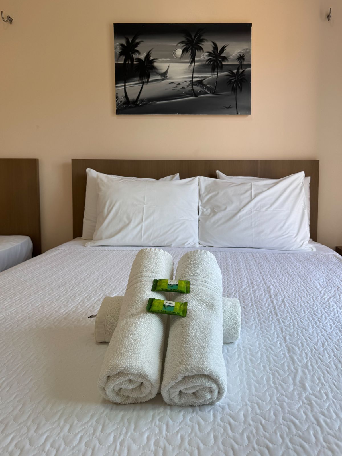 Un lit bien fait, avec du linge de lit blanc et des serviettes pliées, un tableau représentant une scène de plage accroché au mur et une tête de lit en bois.