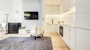 Una cocina moderna con armarios blancos, horno empotrado y microondas. En el suelo hay una alfombra gris y una silla blanca en la sala de estar.