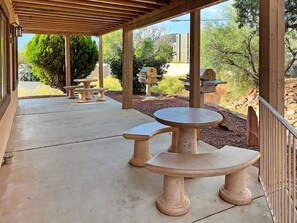 A covered patio area with stone tables and benches, surrounded by greenery and a view of a hilly landscape.