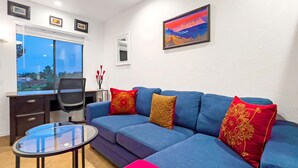 Una sala de estar moderna con un sofá azul, una mesa de centro de vidrio y un escritorio con una silla. Se ve una ventana con vistas a árboles y un cuadro en la pared.