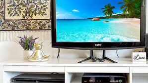 Un televisor de pantalla plana sobre un mueble blanco con un panorama de playa en la pantalla, una pieza decorativa en la pared y una figurita.