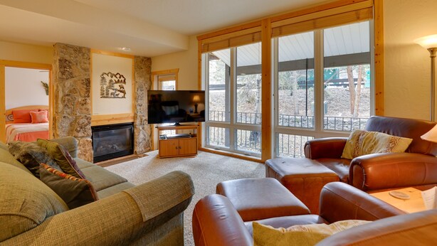 A cozy living room with a stone fireplace, leather sofas, and large windows offering a view of the outdoors. - 1/2 Mi to Granby Ranch: Townhome w/ Fireplace! (Granby)