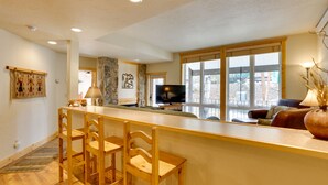 A spacious living area with a wooden bar, seating, and a large window offering a view. - 1/2 Mi to Granby Ranch: Townhome w/ Fireplace! (Granby)