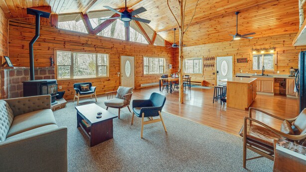 Een ruim houten blokhutinterieur met een hoge plafond, houten wanden, een centraal gelegen openhaard en een eethoek.